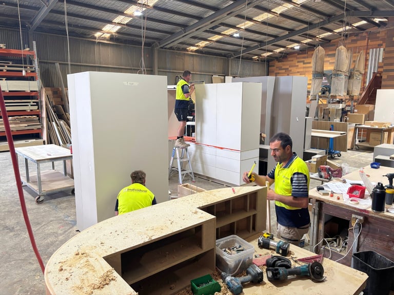 Construction workers in yellow safety vests installing white paneling on a cabinet structure in an industrial warehouse workshop
