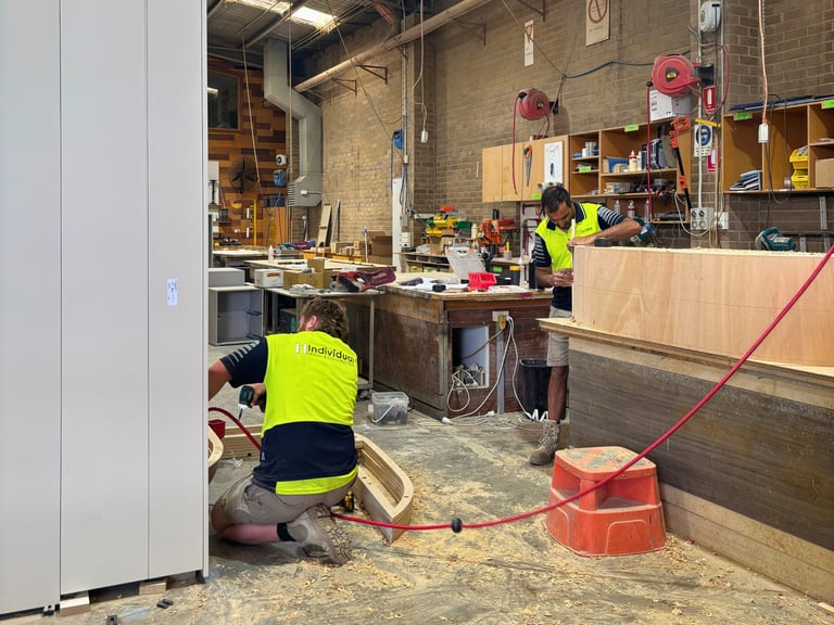Workers in safety vests in a construction workshop, one operating woodworking equipment, with red tools and lumber visible